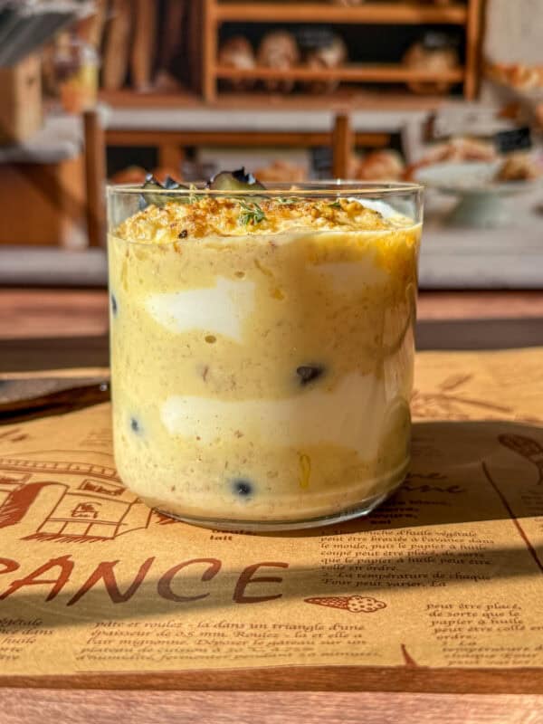 A glass filled with layers of creamy yellow and white yogurt, some blueberries, and a dusting of spices on top, placed on a brown placemat with bakery shelves visible in the blurred background.