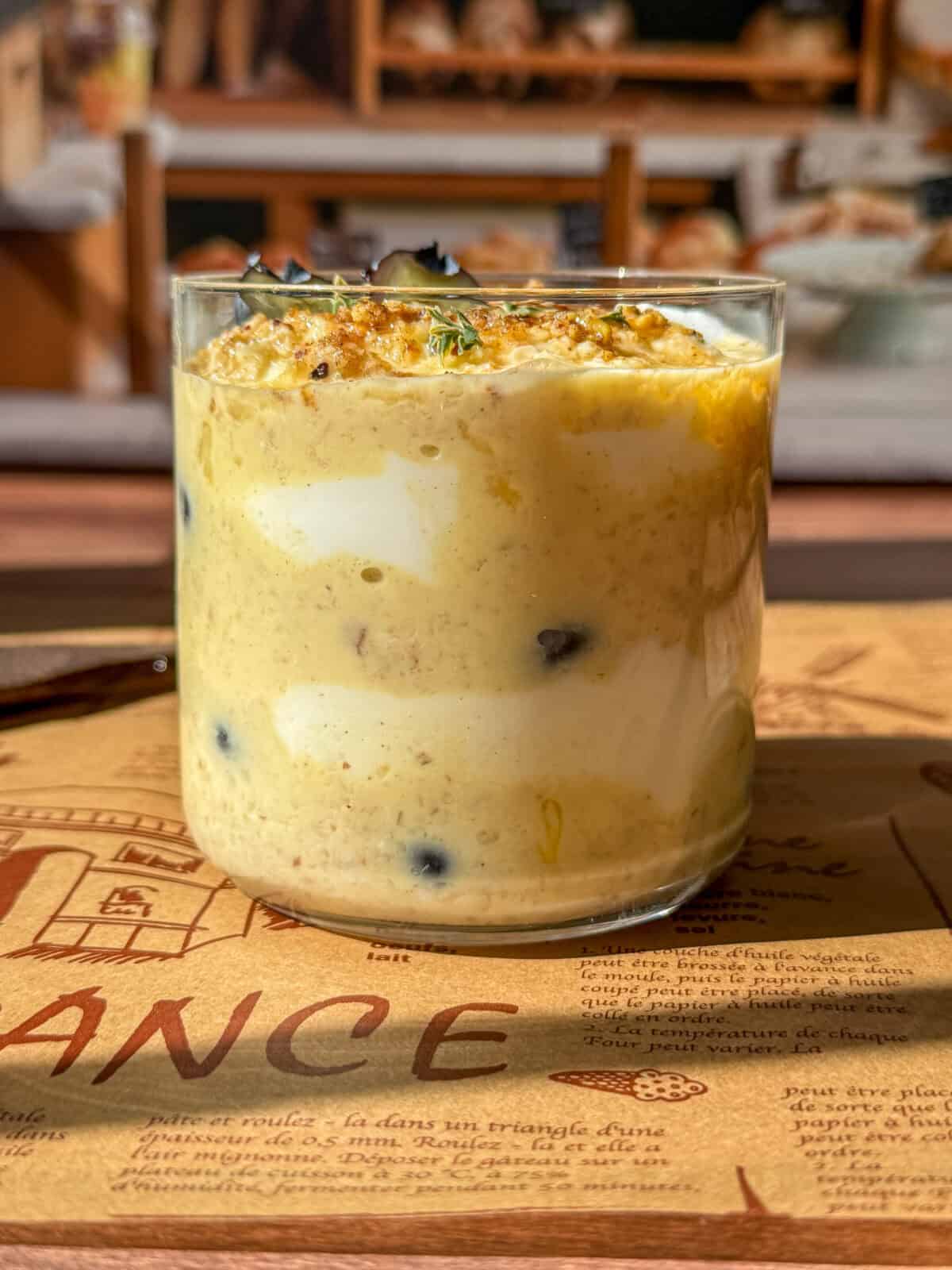 A glass filled with layers of creamy overnight oats topped with granola and herbs, placed on a decorative placemat with bakery shelves blurred in the background.
