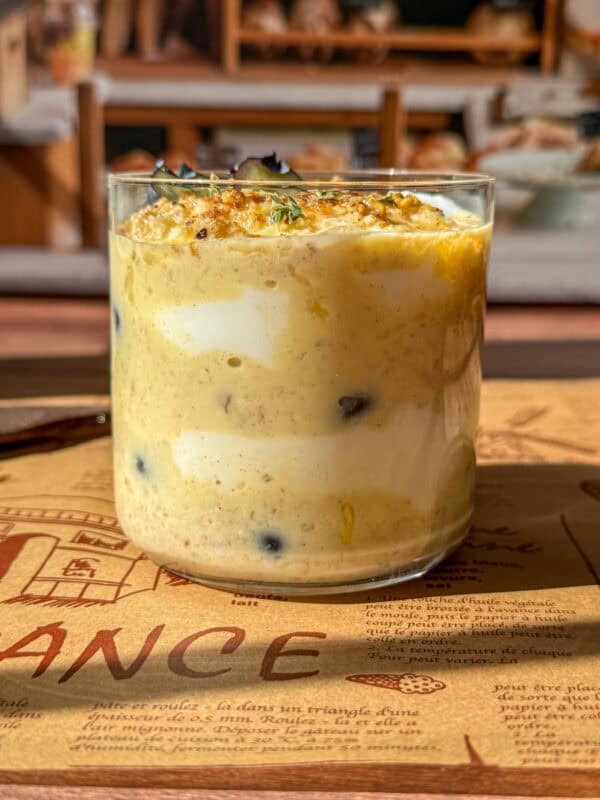 A glass filled with layers of creamy yogurt, yellow pudding, and blueberries, topped with granola and herbs, placed on a decorative placemat with bakery shelves blurred in the background.