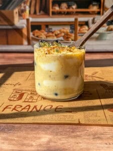 A glass jar filled with creamy, layered dessert topped with blueberries and crumbs, placed on a brown placemat with "FRANCE" written on it. A spoon is in the jar, and a bakery display is visible in the background.