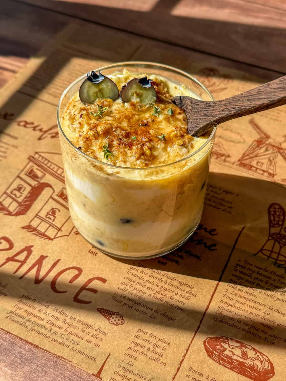 A glass of creamy dessert topped with two blueberries, caramelized sugar, and herbs, sits on a wooden table with a rustic French-themed placemat. A wooden spoon rests in the dessert. Sunlight casts shadows on the scene.