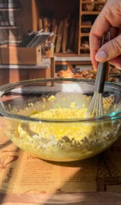 A hand holding a whisk mixes oats and liquid in a glass bowl on a table, with a bakery display of bread and pastries in the background.