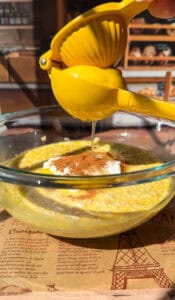 A hand squeezes a yellow lemon squeezer over a glass bowl containing oats, yogurt, and cinnamon, with sunlight illuminating the ingredients and a bakery setting in the background.