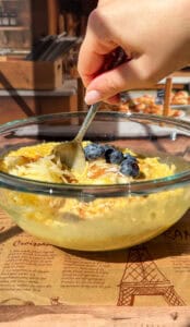 A hand holds a spoon over a glass bowl of oatmeal topped with blueberries, grated apple, and a sprinkle of cinnamon, placed on a paper with Eiffel Tower illustrations.