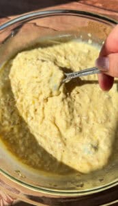 A hand holds a spoon scooping up a thick, yellowish, creamy mixture with visible grains from a glass bowl, lit by natural sunlight.