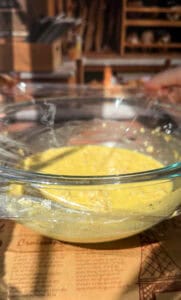A glass bowl filled with yellow batter, covered with plastic wrap, sits on a table in a warmly lit kitchen. The table has a printed recipe sheet beneath the bowl. Shelves and kitchen items are blurred in the background.