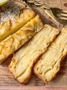 Close-up of a sliced cake with visible layers of thin apple slices, resembling the popular Invisible Pear Cake. The dessert sits on patterned parchment paper, with a fork and cake server in the background.