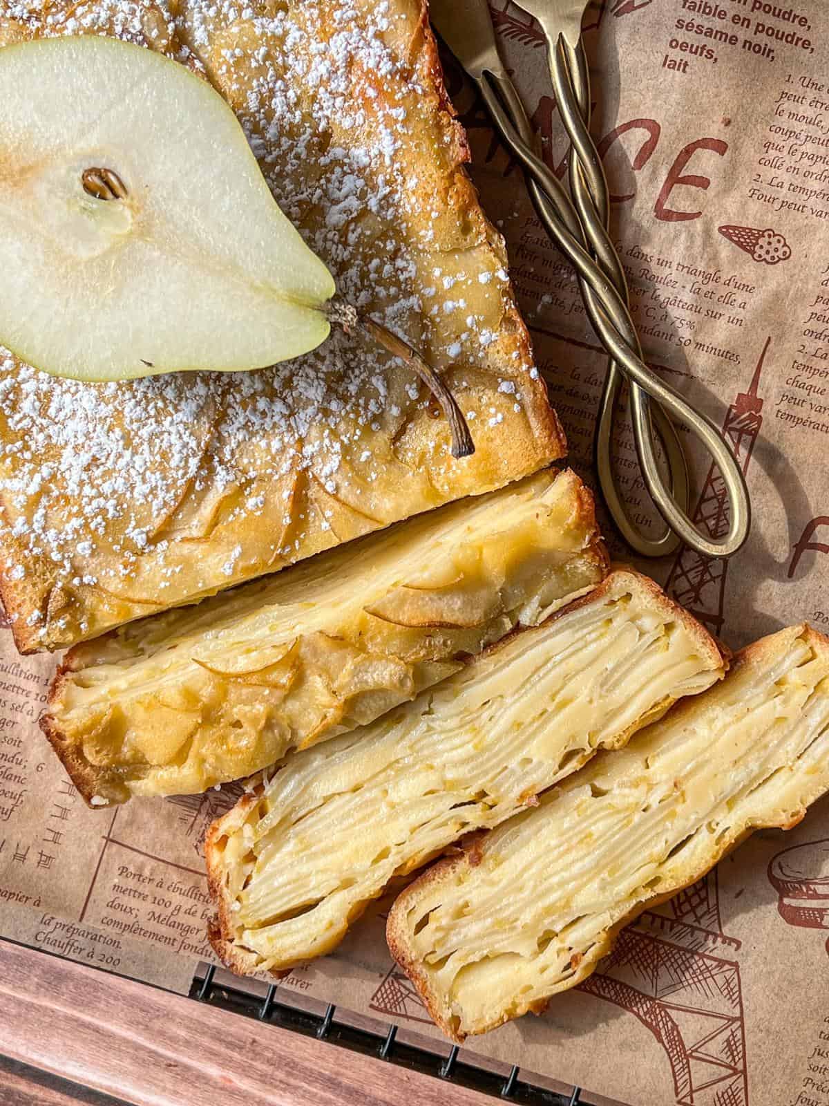 An Invisible Pear Cake dusted with powdered sugar is displayed on parchment paper, topped with a fresh pear slice. Golden cake layers and thinly sliced pears are visible inside, with utensils nearby.