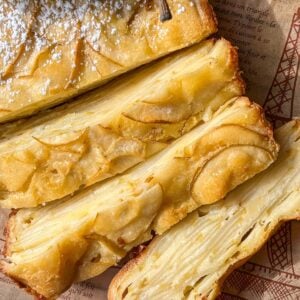 Close-up of several slices of a golden, layered Invisible Pear Cake with thin apple slices visible throughout. The cake is dusted with powdered sugar and placed on a sheet of printed parchment paper.