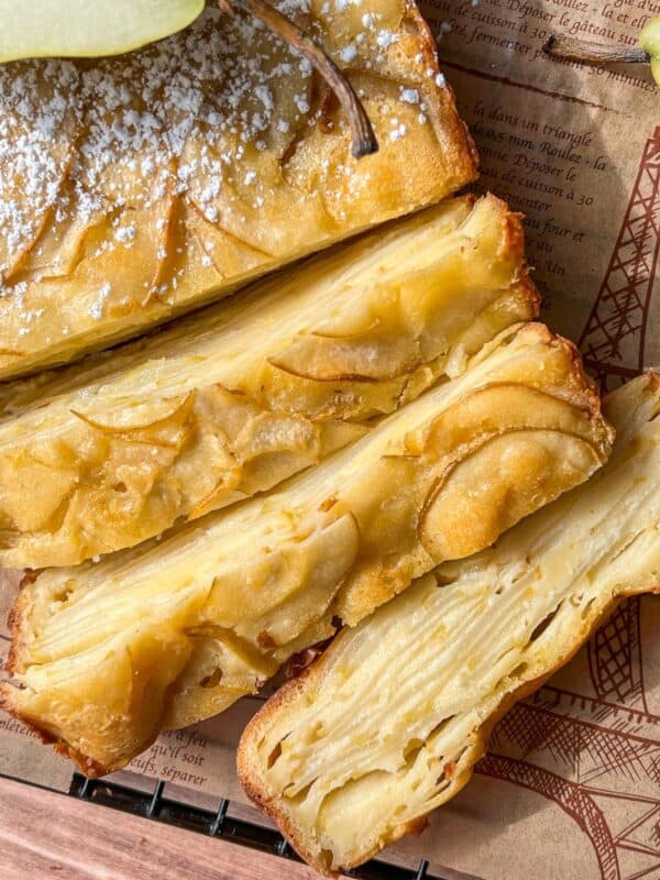 Close-up of several slices of a golden, layered Invisible Pear Cake with thin apple slices visible throughout. The cake is dusted with powdered sugar and placed on a sheet of printed parchment paper.