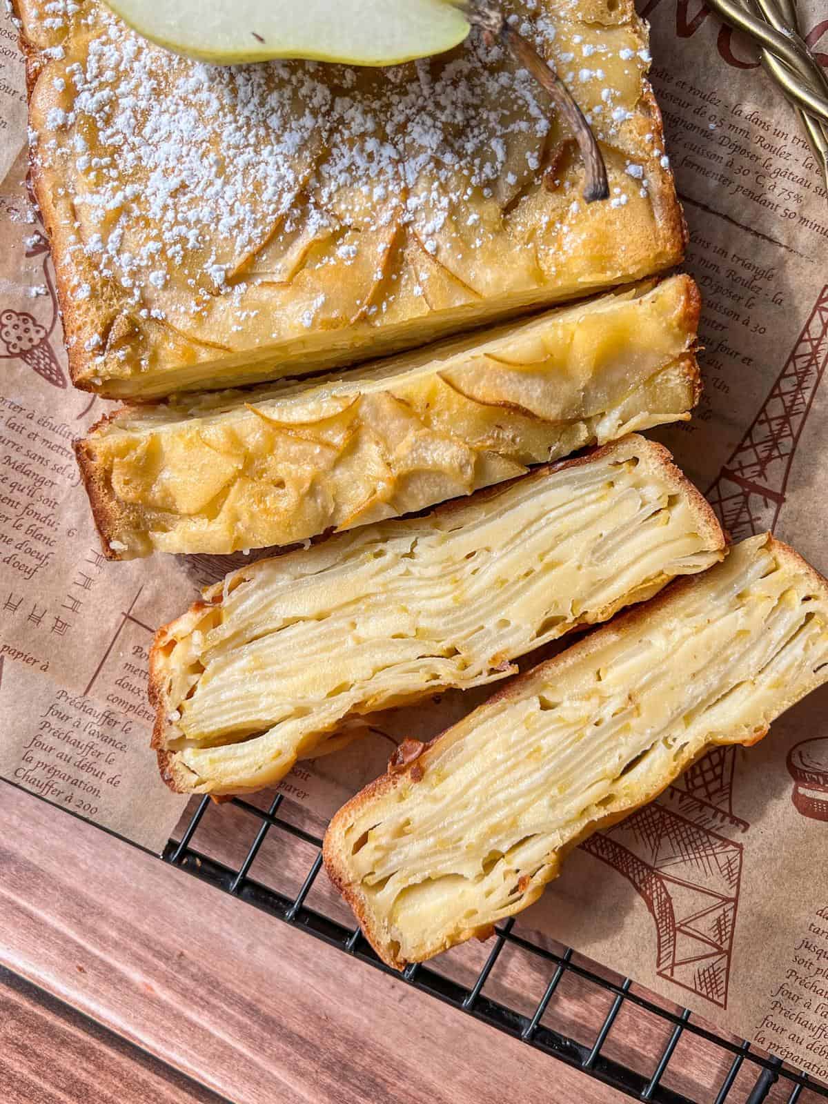 Close-up of a sliced Invisible Pear Cake with powdered sugar on top, revealing thin, layered fruit slices inside. The cake rests on printed parchment paper, set on a cooling rack over a wooden surface.