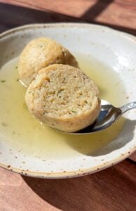 A bowl of clear broth with two sliced matzo balls, one resting on a spoon, sits on a wooden surface in bright sunlight—capturing the essence of Authentic Matzo Ball Soup.