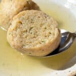 A close-up of a bowl of Authentic Matzo Ball Soup with clear broth and two matzo balls, one being lifted by a spoon. The fluffy texture and visible herbs highlight the traditional comfort of this classic dish.