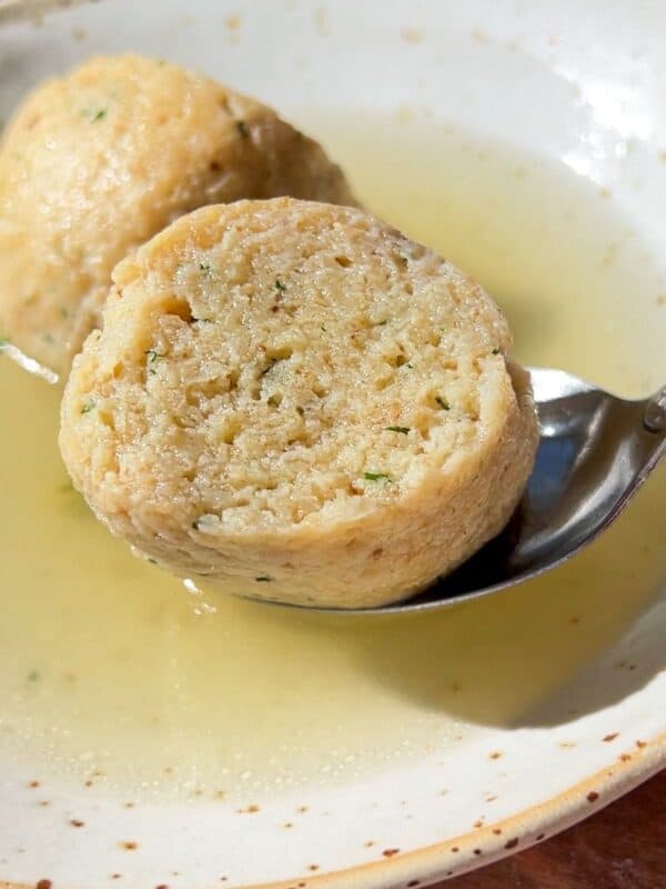 A close-up of a bowl of Authentic Matzo Ball Soup with clear broth and two matzo balls, one being lifted by a spoon. The fluffy texture and visible herbs highlight the traditional comfort of this classic dish.