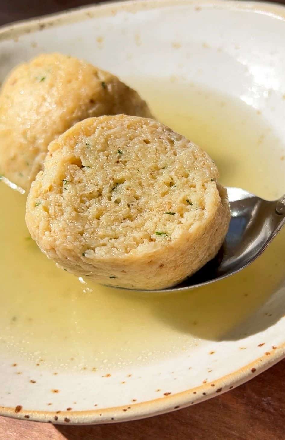 A close-up of Authentic Matzo Ball Soup shows two round matzo balls in clear broth, with one being lifted by a spoon from a shallow, speckled ceramic bowl.