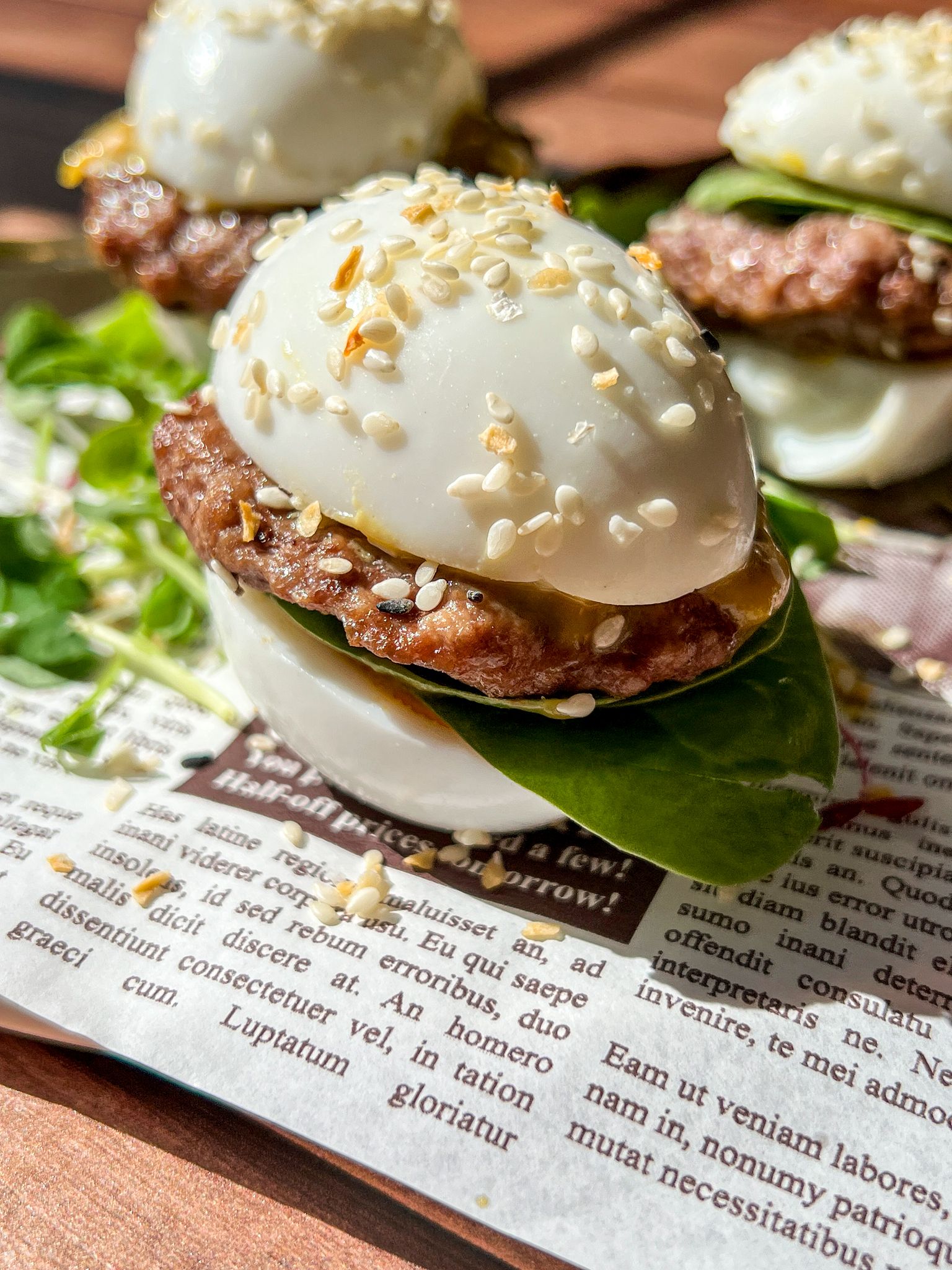 Close-up of Mini Egg Burgers—sliders made with boiled egg halves as buns, filled with a small beef patty, spinach leaves, and sesame seeds sprinkled on top—all placed on a sheet of printed newspaper.