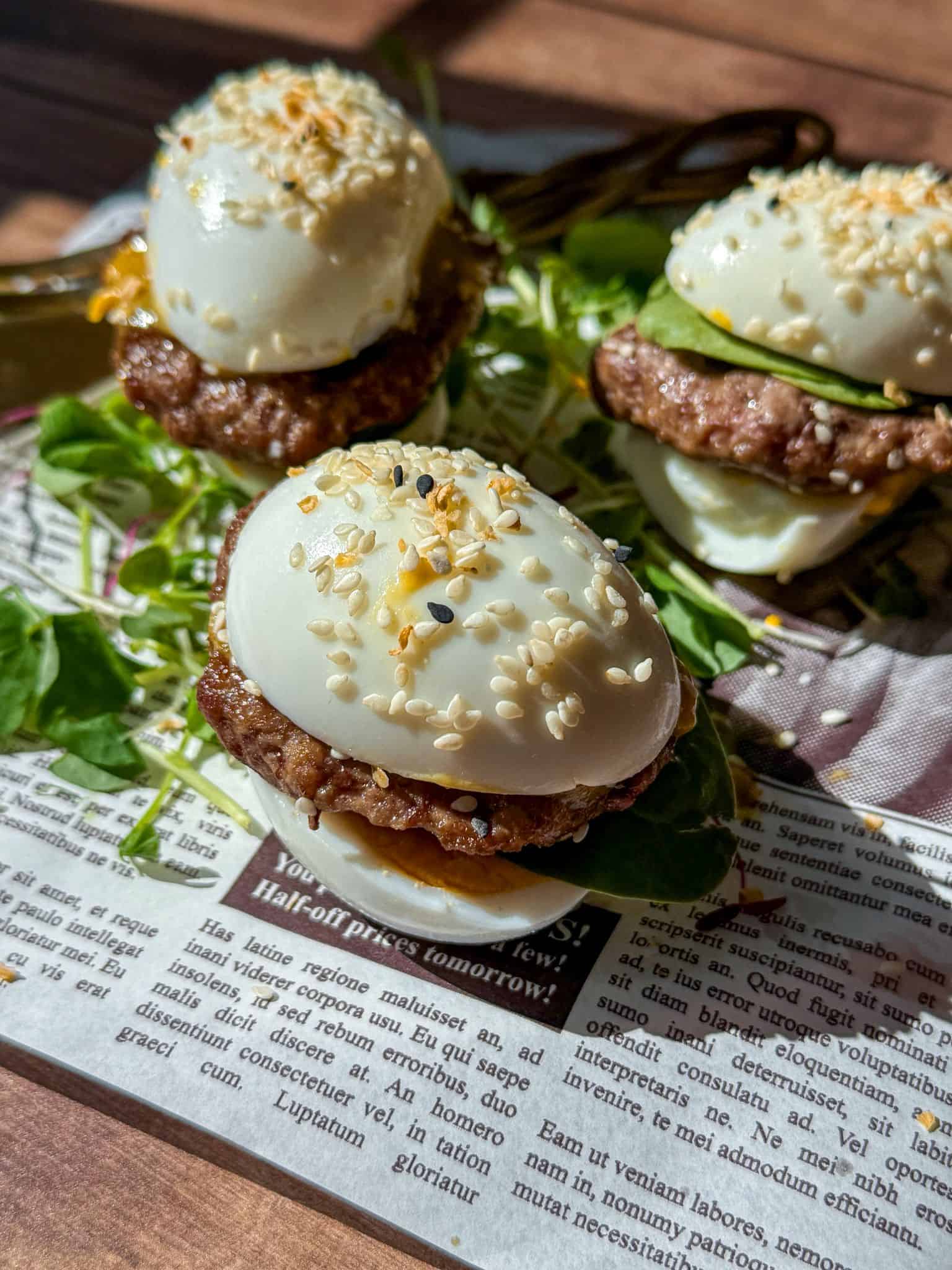 Three Mini Egg Burgers with sesame seeds, burger patties, and greens are arranged on a piece of newspaper under natural sunlight. The eggs act as the “bun,” holding the delicious fillings together.