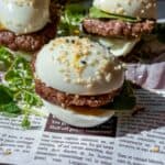 Three slider-style Mini Egg Burgers with hard-boiled eggs as buns, sausage patties, cucumber slices, and sesame seeds, served on fresh greens over a newspaper-patterned surface.