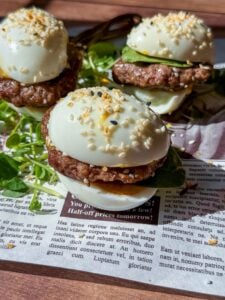 Three slider-style Mini Egg Burgers with hard-boiled eggs as buns, sausage patties, cucumber slices, and sesame seeds, served on fresh greens over a newspaper-patterned surface.