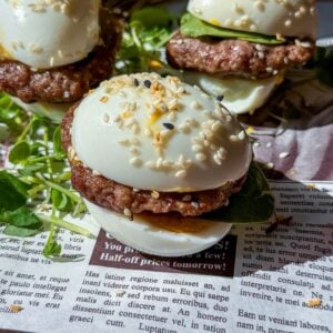 Three slider-style Mini Egg Burgers with hard-boiled eggs as buns, sausage patties, cucumber slices, and sesame seeds, served on fresh greens over a newspaper-patterned surface.