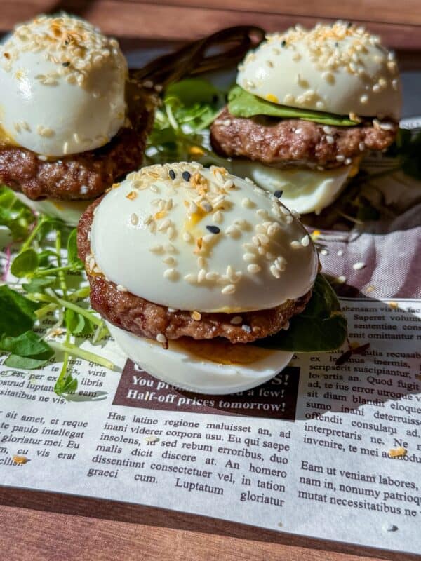 Three slider-style Mini Egg Burgers with hard-boiled eggs as buns, sausage patties, cucumber slices, and sesame seeds, served on fresh greens over a newspaper-patterned surface.