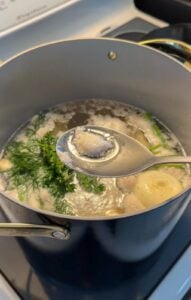 A pot of Easy Homemade Chicken Broth with herbs, onions, and chicken cooks on a stove. A spoon holds grayish foam skimmed from the bubbling surface of the savory liquid.