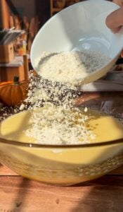 A hand pours flour from a white bowl into a glass mixing bowl of yellow batter. Sunlight streams onto the wooden table, hinting at an Invisible Pear Cake in progress, while a pumpkin sits blurred in the background.