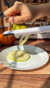 A hand slices a green pear using a white mandoline slicer over a white plate, preparing thin pear slices for Invisible Pear Cake. A small pumpkin sits in the background on a wooden surface.