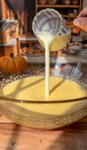 A hand pours creamy liquid from a glass measuring cup into a large bowl of yellow batter on a wooden table, preparing an Invisible Pear Cake, with baked goods and a pumpkin in the kitchen background.