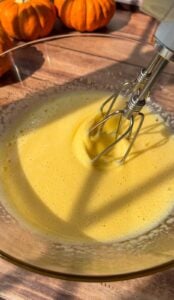 A close-up of an electric mixer blending yellow Invisible Pear Cake batter in a glass bowl on a wooden surface, with small pumpkins in the background and sunlight casting shadows across the scene.
