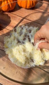 A hand mixes a yellow and white powdered mixture in a glass bowl on a wooden surface, with small orange pumpkins in the background—prepping ingredients for an Invisible Pear Cake.