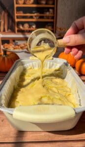 A hand pours creamy batter from a glass cup over thinly sliced apples arranged in a baking dish, creating an Invisible Pear Cake. Small pumpkins and bread are visible in the warm, autumn-themed background.