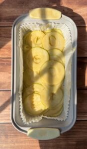 A rectangular baking pan lined with parchment paper holds thinly sliced green apples, arranged in overlapping layers for an Invisible Pear Cake. Sunlight casts gentle shadows on the wooden surface beneath the pan.