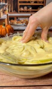A hand mixes thinly sliced green apples in batter for an Invisible Pear Cake inside a glass bowl, with a small pumpkin and bakery shelves filled with bread visible in the background.