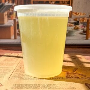 A clear plastic container filled with light yellow liquid sits on a brown paper mat in a bakery setting, with shelves of bread and pastries blurred in the background.