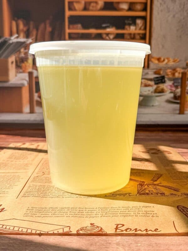 A clear plastic container filled with light yellow liquid sits on a brown paper mat in a bakery setting, with shelves of bread and pastries blurred in the background.