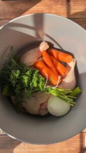 A pot filled with raw chicken, whole carrots, celery stalks, fresh dill, parsley, and a peeled white onion sits on a wooden surface in natural sunlight—perfect for making Easy Homemade Chicken Broth.