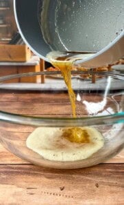 Melted butter is being poured from a saucepan into a clear glass mixing bowl on a wooden surface.