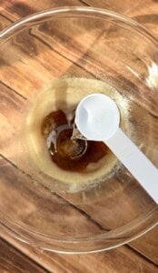 A clear glass bowl on a wooden surface contains a brown liquid. A white measuring spoon pours a clear liquid, possibly water or vanilla extract, into the bowl.