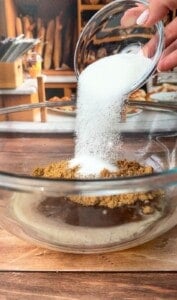 A hand pours white granulated sugar from a small glass bowl into a larger glass bowl containing brown sugar, with a kitchen setting and bread in the background.