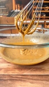 A whisk covered in thick, creamy batter is held above a glass bowl filled with more batter, set on a wooden surface, with shelves of bread and kitchen items blurred in the background.
