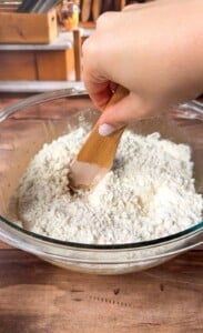 A hand holding a wooden spatula mixes flour in a clear glass bowl on a wooden surface, with kitchen items blurred in the background.