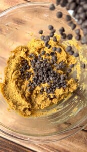 A glass bowl filled with cookie dough sits on a wooden surface. Chocolate chips are being poured into the dough from above.