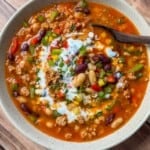 A bowl of hearty chili topped with sour cream, diced green onions, and red bell peppers, featuring kidney beans, white beans, and ground meat in a rich tomato-based broth, with a wooden spoon on the side.