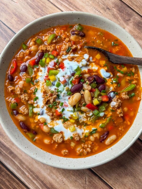 A bowl of hearty chili topped with sour cream, diced green onions, and red bell peppers, featuring kidney beans, white beans, and ground meat in a rich tomato-based broth, with a wooden spoon on the side.