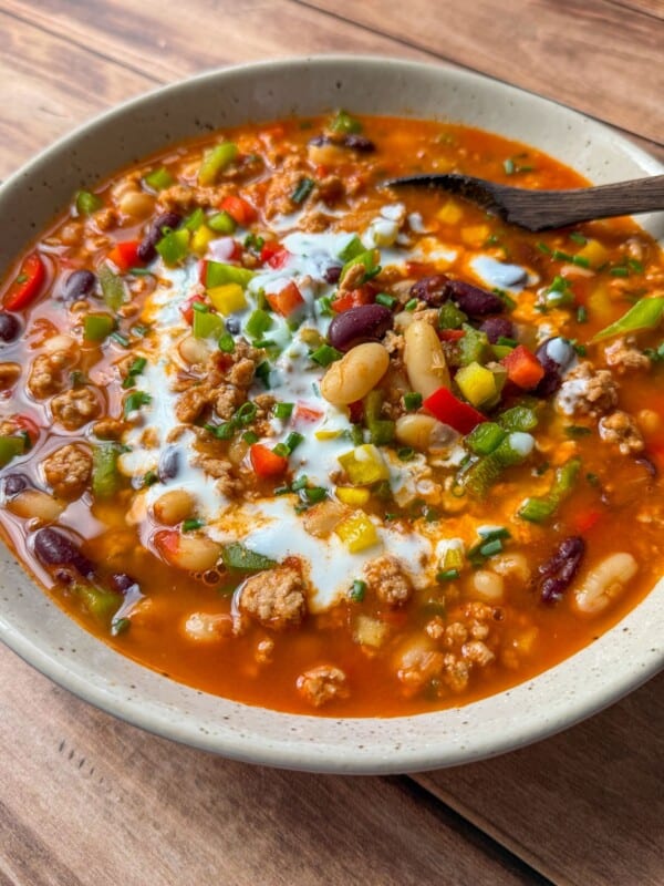 A bowl of hearty chili with ground meat, red and white beans, chopped peppers, green onions, and a swirl of sour cream on top, served with a wooden spoon on a wooden table.