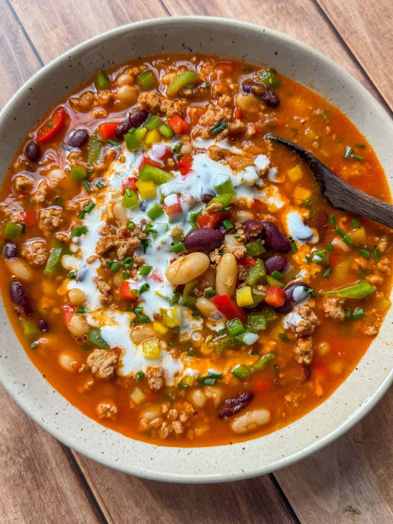 A bowl of chili with ground meat, kidney beans, white beans, red and green bell peppers, topped with sour cream and chopped chives, with a wooden spoon in the bowl on a wooden surface.