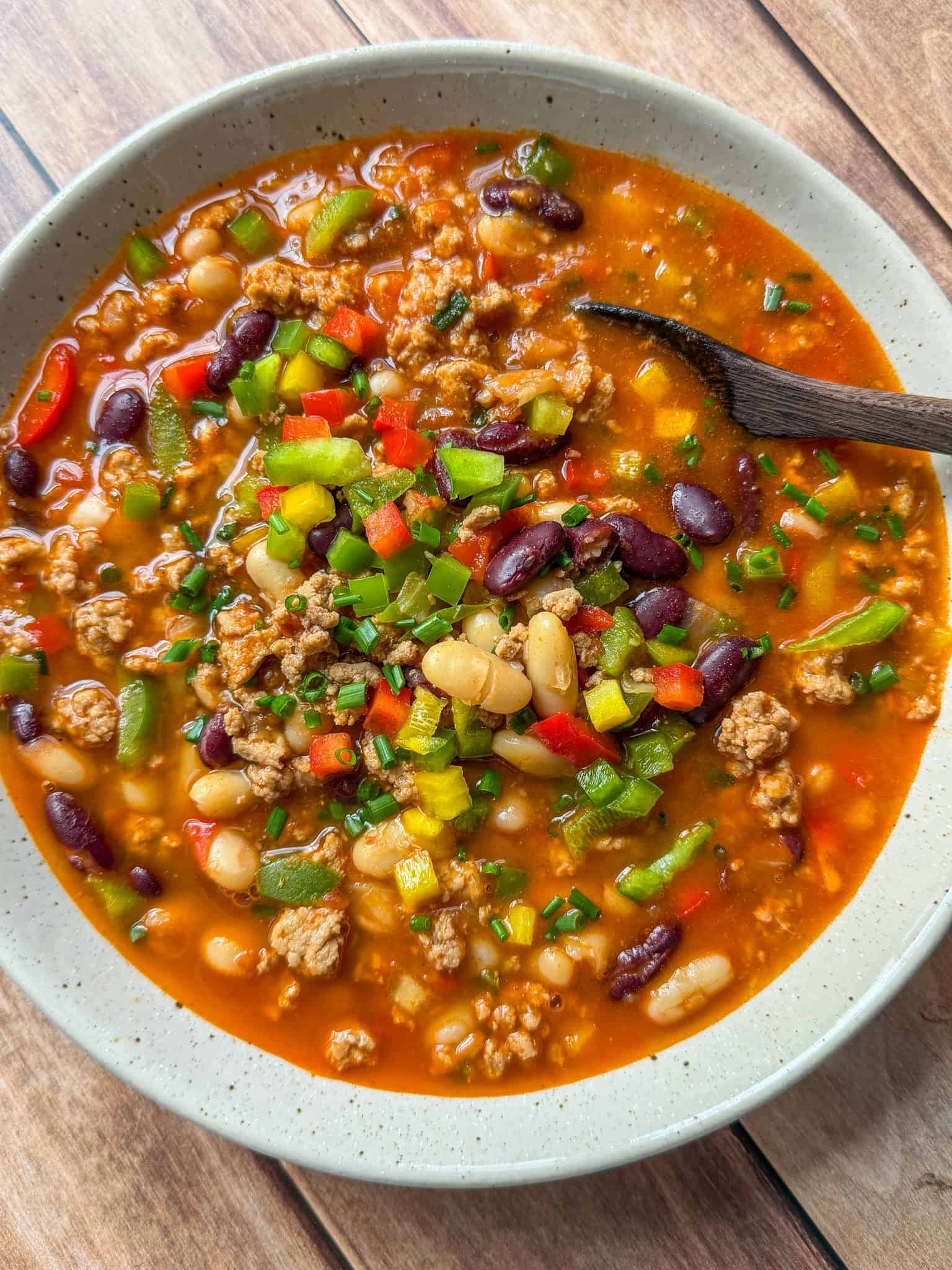 A bowl of chili with ground meat, kidney beans, white beans, diced green and red bell peppers, topped with chopped chives, and a wooden spoon resting inside the bowl on a wooden surface.