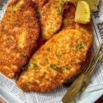 A plate of golden, crispy breaded schnitzels garnished with chopped parsley, served with lemon wedges and a fork on a piece of newspaper-style parchment paper.