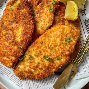 A plate of golden, crispy breaded schnitzels garnished with chopped parsley, served with lemon wedges and a fork on a piece of newspaper-style parchment paper.